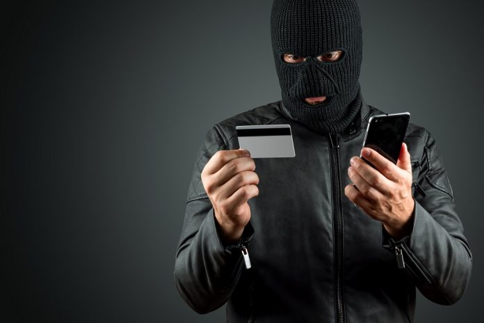 Robber, a thug in a balaclava holds a credit card in his hands on a dark background. Robbery, hacker, crime, theft. Copy space. phishing nel settore finanziario: è boom di credential stuffing